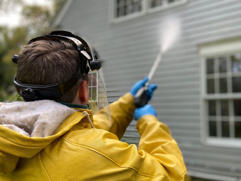 Technician in a yellow rain jacket and face shield pressure washing gray vinyl siding on a house