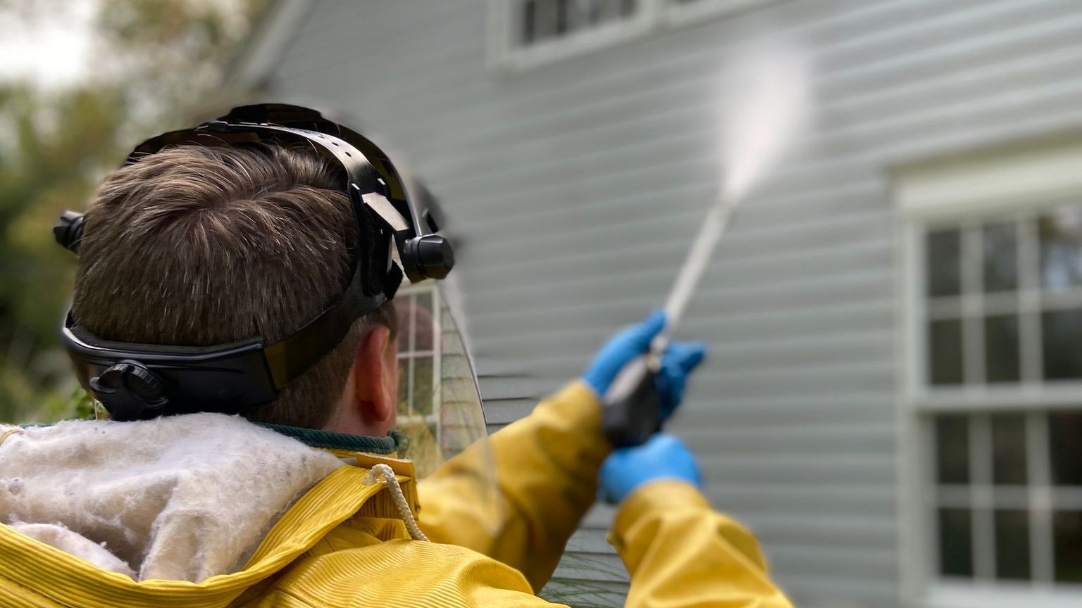 Technician in a yellow rain jacket and face shield pressure washing gray vinyl siding on a house